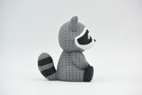 Knitted raccoon decorative figure placed on a wooden shelf beside books and a small plant